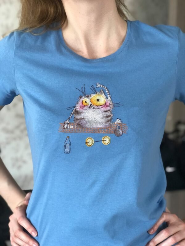 Cross stitch embroidery of a cute yoga cat sitting on a mat with small props and bright colors, displayed on t-shirt