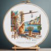 131 Cross stitch embroidery of a ginger cat sitting on a windowsill with a seaside town view, books, plants, and warm summer colors, displayed in a wooden hoop