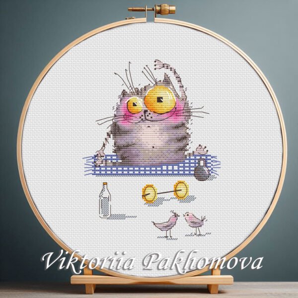 Cross stitch embroidery of a cute yoga cat sitting on a mat with small props and bright colors, displayed in a wooden hoop