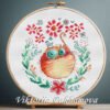 Cross stitch embroidery of a ginger cat inside a floral wreath with bright flowers and green leaves, displayed in a wooden hoop