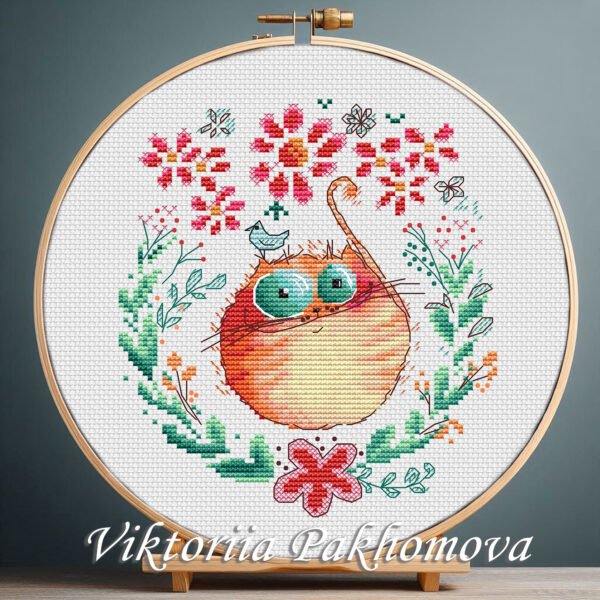 Cross stitch embroidery of a ginger cat inside a floral wreath with bright flowers and green leaves, displayed in a wooden hoop