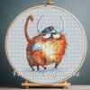 Cross stitch embroidery of a whimsical ginger cat dressed as a Viking with a horned helmet and armor, displayed in a wooden hoop