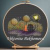 Cross stitch embroidery of a black and ginger cat sitting together under glowing trees with soft romantic details, displayed in a wooden hoop