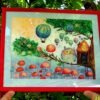 Framed cross stitch artwork of a whimsical owl beside a tiny forest house with colorful mushrooms and balloons
