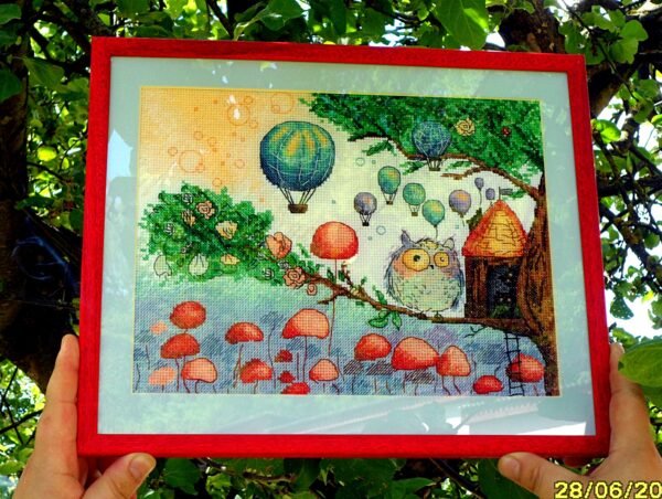 Framed cross stitch artwork of a whimsical owl beside a tiny forest house with colorful mushrooms and balloons