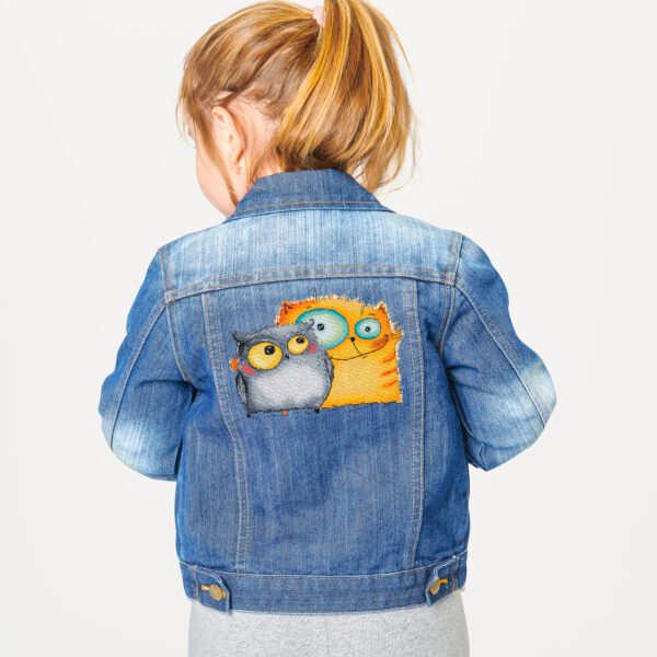 Cross stitch embroidery of a cheerful cat and owl standing together with bright colors and playful details, displayed on a denim jacket