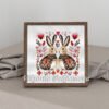 Framed cross stitch artwork of two romantic folk‑art bunnies surrounded by Scandinavian floral motifs