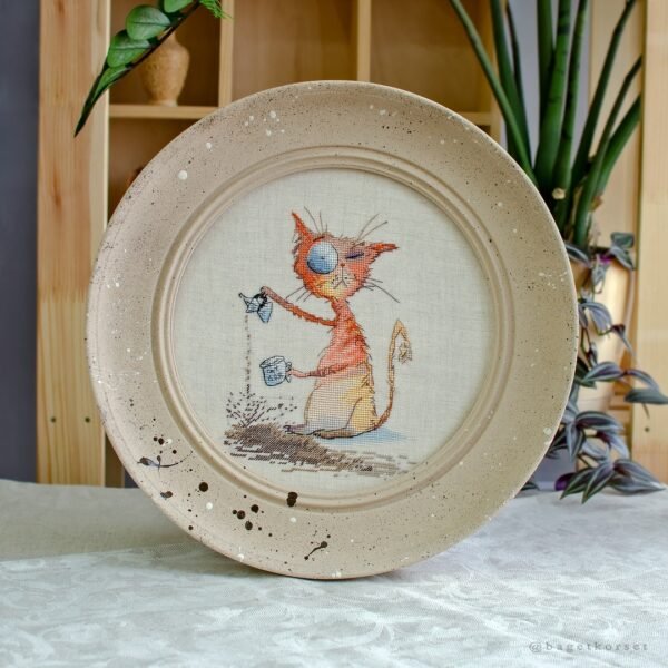 Framed cross stitch artwork featuring a whimsical ginger cat with a coffee cup and bold colorful shading.