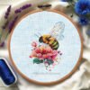 Peony and bee cross stitch pattern in a wooden embroidery hoop, detailed botanical floral design in pink and yellow tones