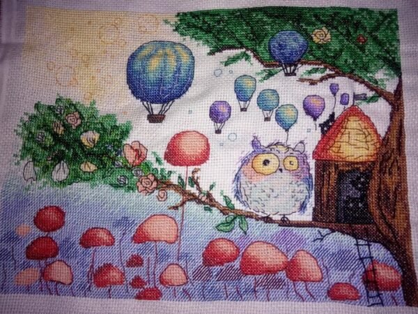Framed cross stitch artwork of a whimsical owl beside a tiny forest house with colorful mushrooms and balloons