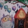 Framed cross stitch artwork of a whimsical owl beside a tiny forest house with colorful mushrooms and balloons