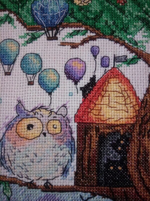 Framed cross stitch artwork of a whimsical owl beside a tiny forest house with colorful mushrooms and balloons