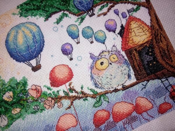 Framed cross stitch artwork of a whimsical owl beside a tiny forest house with colorful mushrooms and balloons