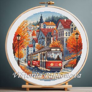 Cross stitch embroidery of an autumn city scene with a red tram moving along a street lined with colorful fall trees and European-style buildings, displayed in a wooden hoop
