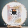 Cross stitch embroidery of a funny black cat posed like a Da Vinci sketch inside a circle and square, with warm background colors, displayed in a wooden hoop