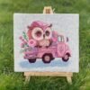 Cross stitch of baby owl in pink truck displayed on small easel outdoors, whimsical embroidery design