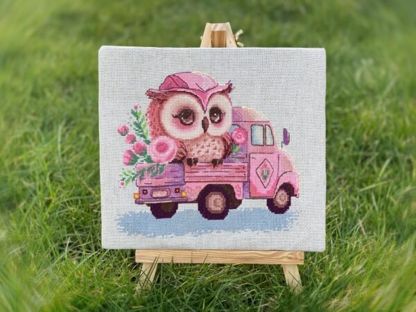 Cross stitch of baby owl in pink truck displayed on small easel outdoors, whimsical embroidery design