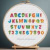 Cross stitch embroidery of a colorful dinosaur alphabet with rainbow Jurassic letters displayed in a wooden hoop