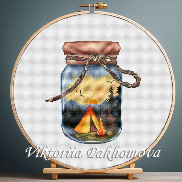 Cross stitch embroidery of a glass jar with a tent, campfire and forest landscape held in a wooden hoop