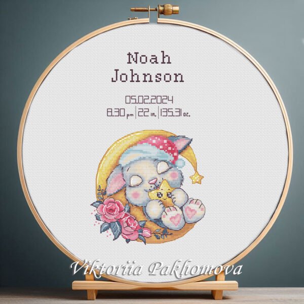 Cross stitch embroidery of a personalized sleeping bunny birth announcement displayed in a wooden hoop with custom newborn details.