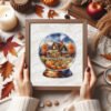 Framed cross stitch artwork showing a glass ball with an autumn landscape, warm trees and a small house