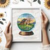 Framed cross stitch artwork showing a glass ball with a spring garden, cozy house and bright floral colors