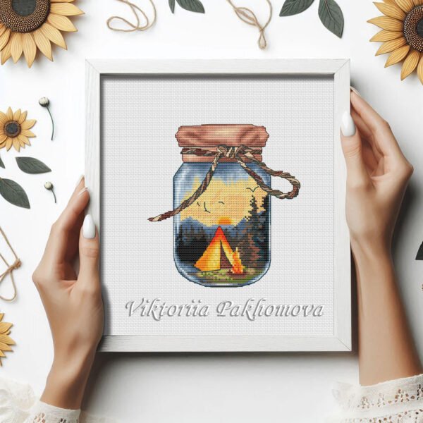 Framed cross stitch of a glass jar camping scene with mountains, trees and a glowing tent.