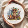 Cross stitch embroidery of a camping owl sitting by a campfire with autumn leaves in a wooden hoop