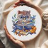 57 Autumn bookworm owl cross stitch pattern in a wooden hoop with pumpkins, books, and warm seasonal colors.