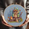 Cross stitch embroidery of a camping owl sitting by a campfire with autumn leaves in a wooden hoop