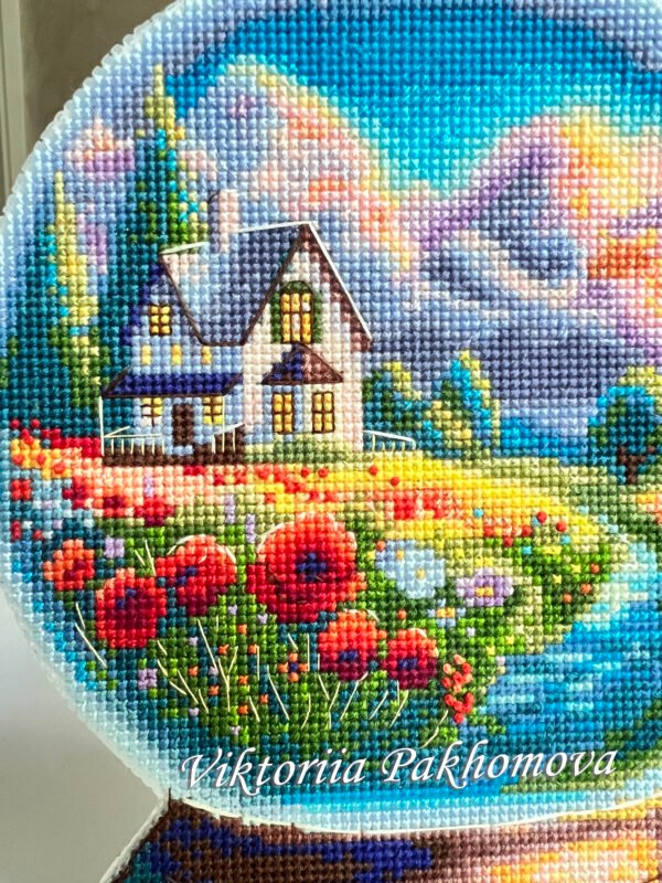 Framed cross stitch artwork showing a glass ball with a poppy field, warm sky and a small house