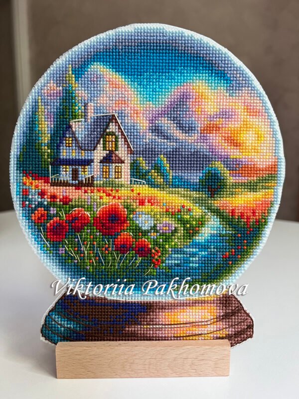 Framed cross stitch artwork showing a glass ball with a poppy field, warm sky and a small house
