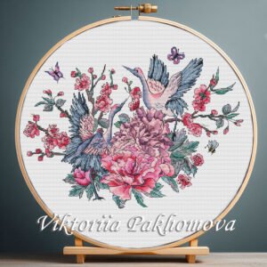 Cross stitch pattern displayed in embroidery hoop showing two cranes surrounded by pink peonies, blossoms and butterflies on white fabric.