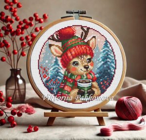 Baby reindeer cross stitch pattern in hoop – cozy woodland animal with festive hat and scarf, holding a cup of cocoa among snowy trees