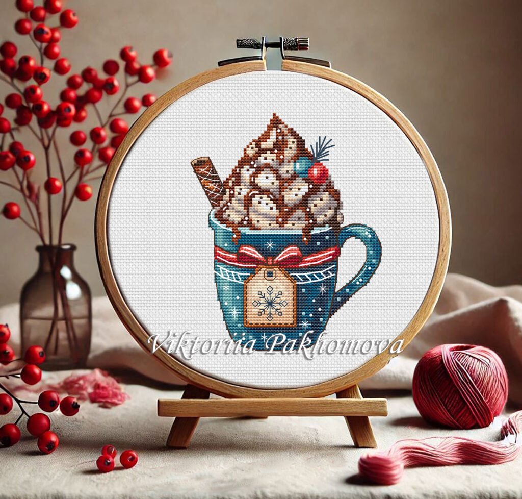 Christmas gingerbread house coffee cup cross stitch pattern in embroidery hoop with pink festive mug, whipped cream, and candy cane, cozy holiday décor design.
